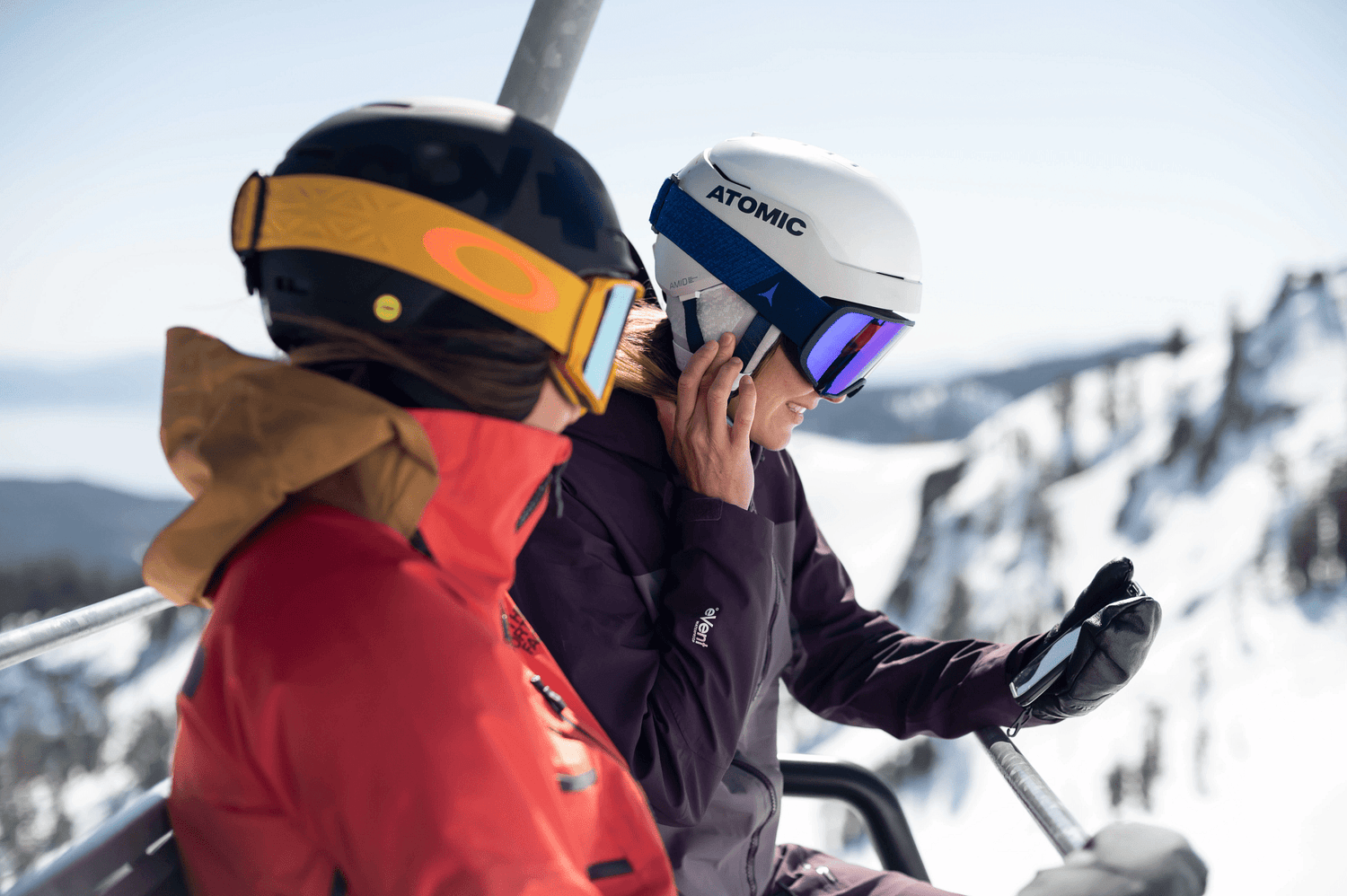 Two skiers on chairlift wearing audio-ready Atomic ski helmets with Aleck speakers, snowy mountain backdrop