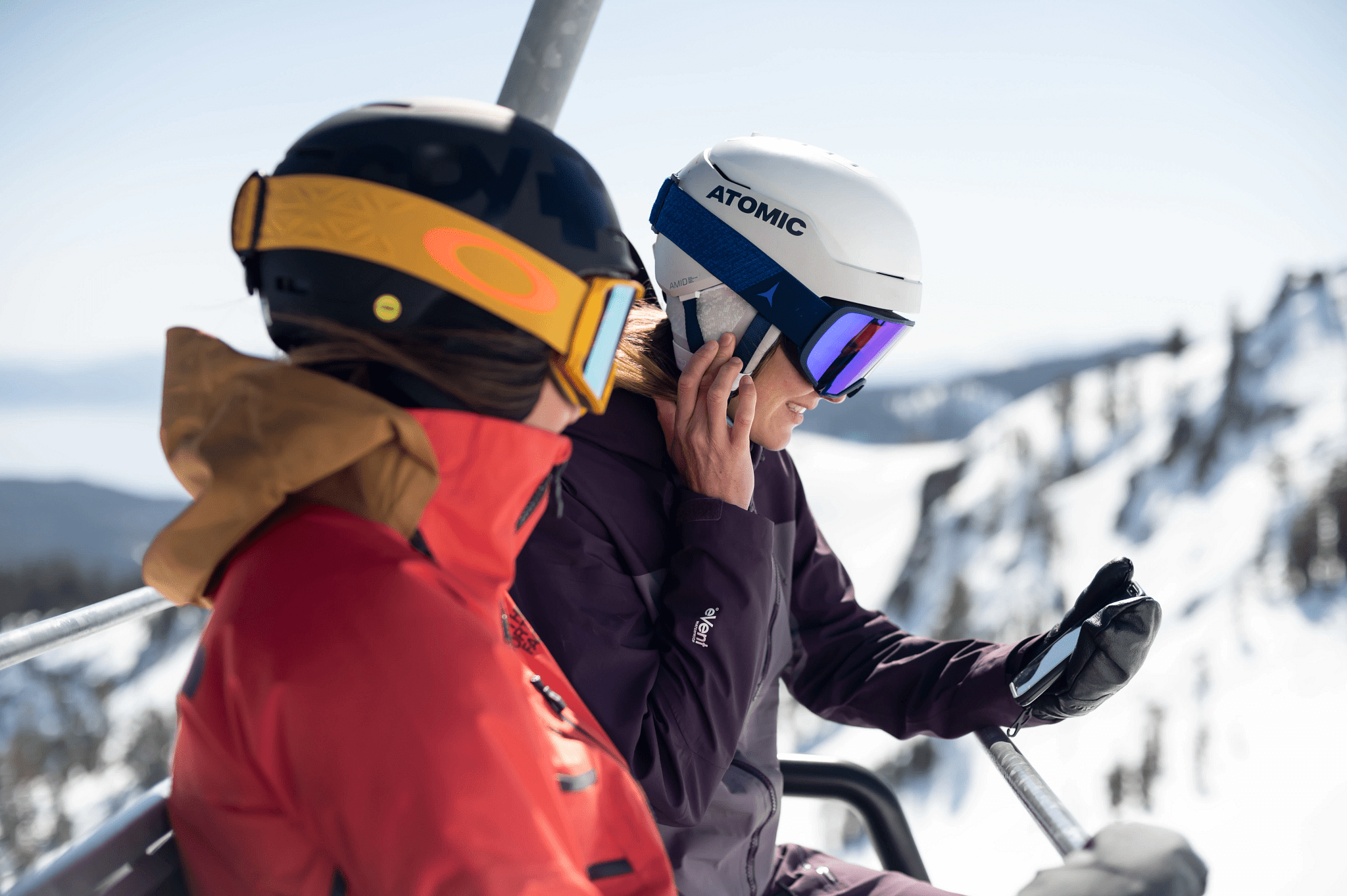 Two skiers on chairlift wearing audio-ready Atomic ski helmets with Aleck speakers, snowy mountain backdrop